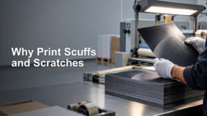 Gloved worker inspects a black printed sheet with visible scratch-like marks over a stack of matching sheets at a finishing table under a bright inspection lamp.
